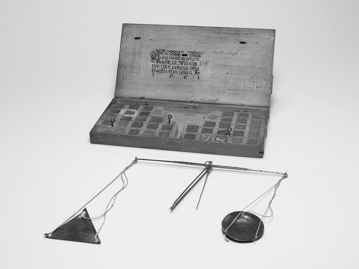 Open wooden box of brass coin weights beside a small hand balance with two pans