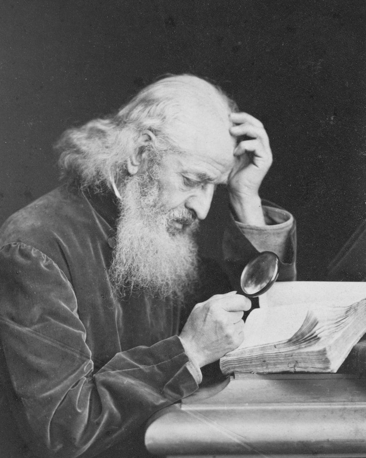 A white-bearded man bent over an open manuscript, reading it through a magnifying glass